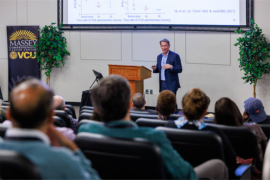 Ned Sharpless speaking holding a clicker beside a podium. In the background, there is a tall black Massey-branded sign